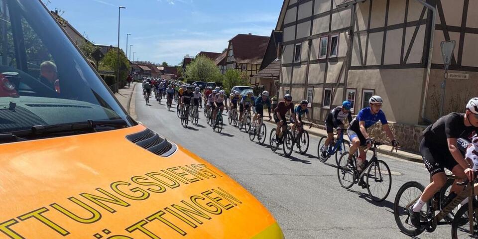 Die Motorhaube eines Rettungswagens ist zu sehen. Das Fahrzeug steht auf einer Straße in einem Ort. Auf der Straße fahren Radsportler. 