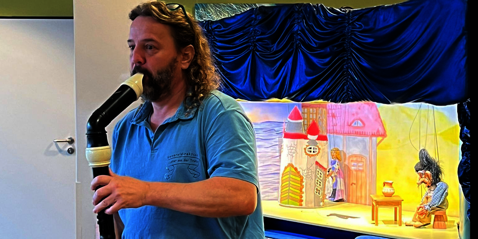 Ein Mann in einem blauen Poloshirt, schulterlange lockige Haar und Bart. Er hat eine Brille in die Haare gesteckt und spielt auf eine schwarzen Bassflöte. . Er steht vor einem Kasten mit blauem Vorhang, der der den Blick auf eine erleuchtete kleine Bühne freigibt.