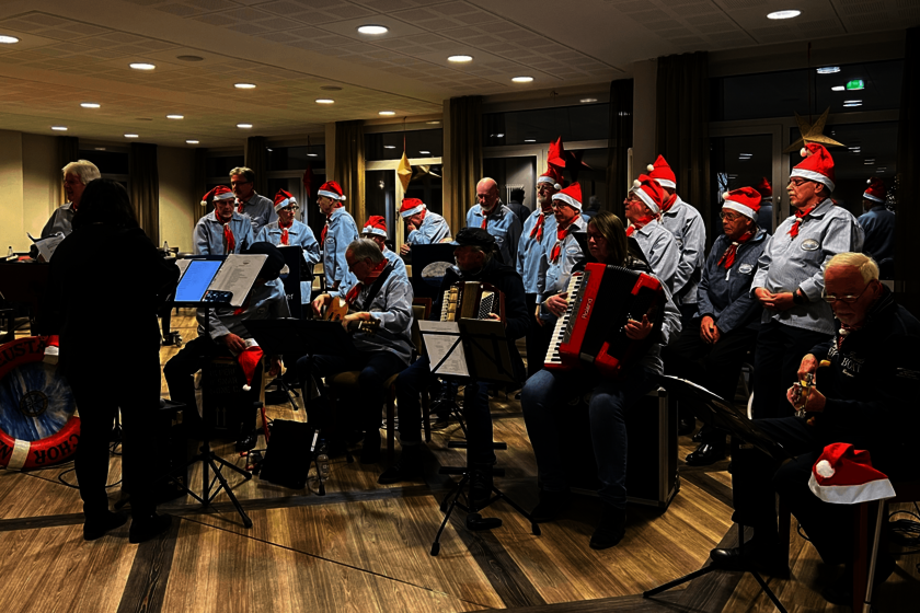 Eine Gruppe von Menschen mit Instumenten hinter Notenständer. Die meisten tragen hellblaue Hemden, roto Holztücher und rote Weihnachts-Zipfelmützen.