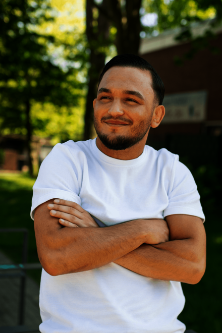 Ein Mann in weißem T‑Shirt steht draußen und verschränkt die Arme