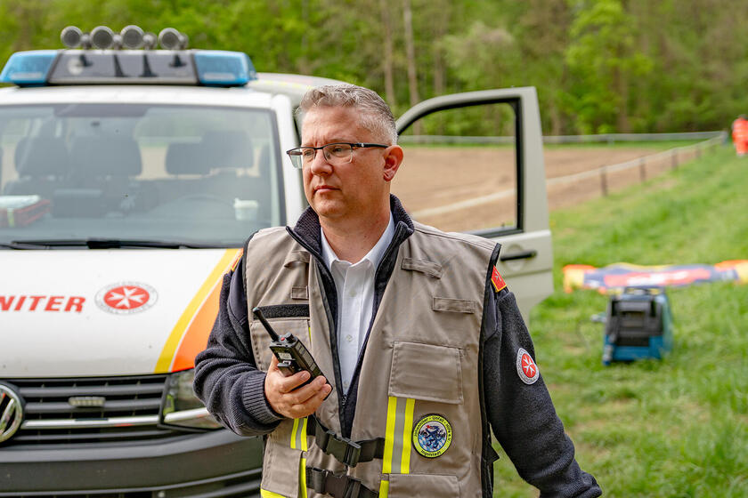 Ein Johanniter-Einsatzleiter mit Funkgerät steht vor einem Einsatzfahrzeug auf einer Wiese und blickt aufmerksam in Richtung Einsatzbereich.