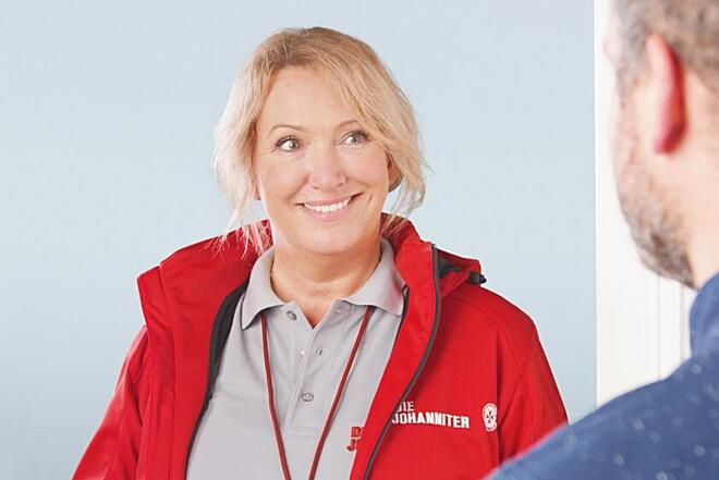 Eine Frau in roter Johanniter-Jacke lächelt einen Mann an, der offenbar gerade die Tür geöffnet hat.
