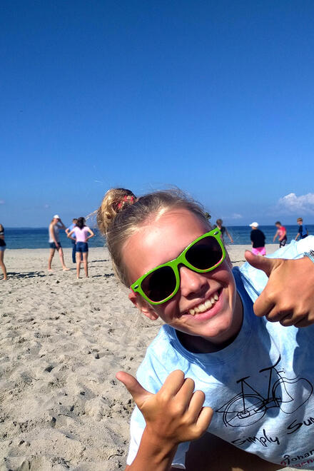 Ein Mädchen mit Sonnenbrille am Strand guckt in die Kamera und zeigt beide Daumen hoch.