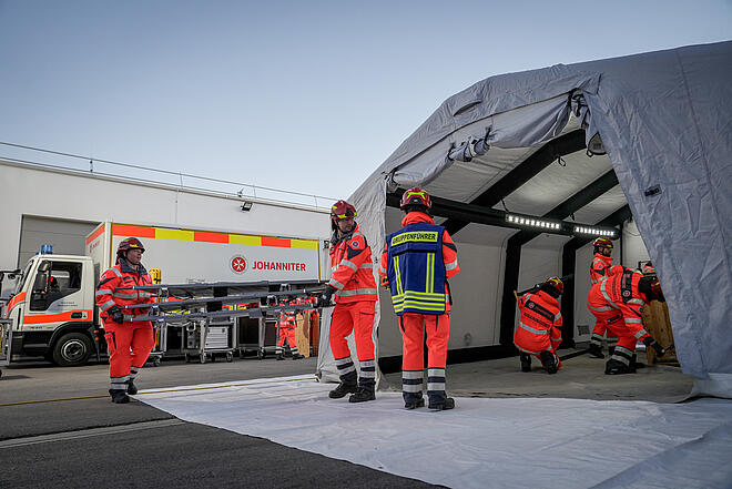 Einsatzkräfte bauen ein Zelt auf und bringen Tragen hinein. Im Hintergrund: Ein Gerätewagen der Johanniter.