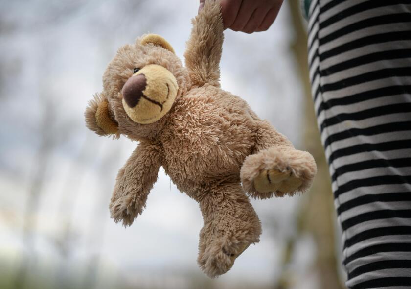 Ein Kind hält einen beigefarbenen Teddybären locker in der Hand, während es in gestreifter Hose danebensteht. 