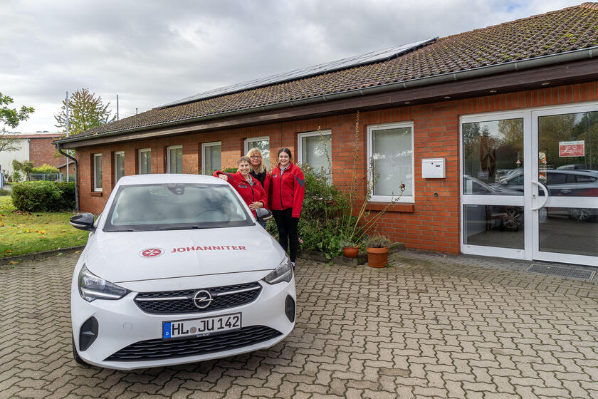 Auf dem Bild sind drei Mitarbeiterinnen des Ambulanten Pflegedienstes zu sehen, die neben einem Auto der Johanniter stehen. Im Hintergrund ist die Dienststelle in Eutin zu sehen, auf dem Dach sind die Solarmodule.