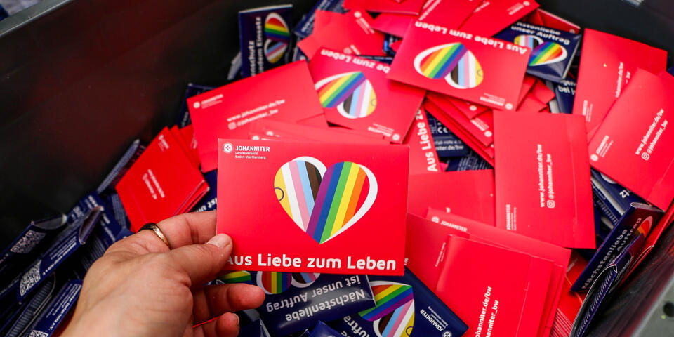 Eine Person hält eine Postkarte der Johanniter in Rot, mit einem Regenbogenherz und der Aufschrift "Aus Liebe zum Leben" und dem Johanniter-Logo. Im Hintergrund ist eine Kiste mit vieler dieser Karten, auch in blau.