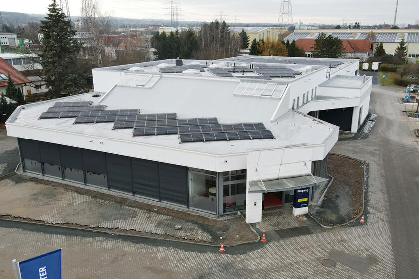Das Gebäude ist von oben aus der Luft fotografiert. Man sieht das weiße Dach mit den Solarpanelen sowie die vordere Fensterfront. 