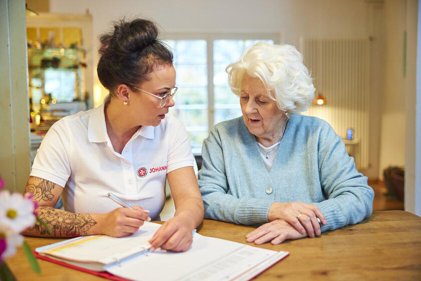 Johanniter Mitarbeiterin bespricht Dokumente mit Seniorin am Küchentisch.