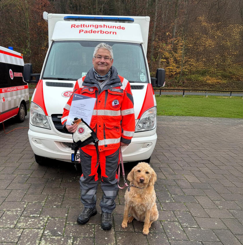 Das Johanniter Rettungshund-Team Ulrich Sabisch und seine Hündin Lucy stehen zufrieden mit einer Urkunde vor einem Rettungswagen in Paderborn. Im Hintergrund sind Büsche und Bäume zu sehen.