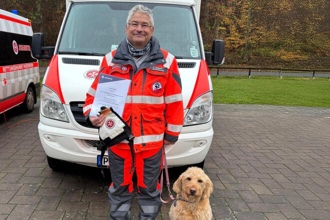 Das Johanniter Rettungshund-Team Ulrich Sabisch und seine Hündin Lucy stehen zufrieden mit einer Urkunde vor einem Rettungswagen in Paderborn. Im Hintergrund sind Büsche und Bäume zu sehen.