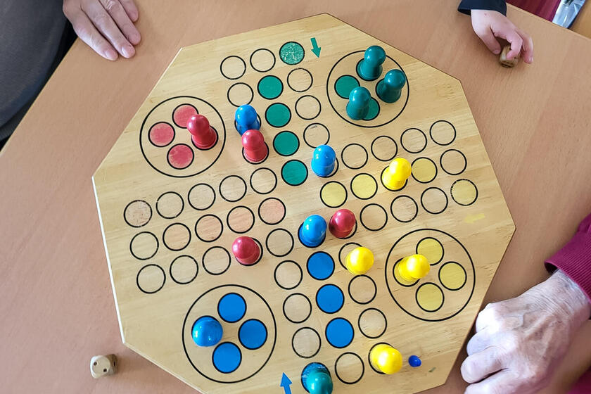 Spielbrett mit bunten Holzspielfiguren (rot, blau, gelb, grün) und Händen mehrerer Personen beim Brettspiel.