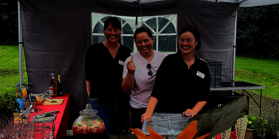 drei lächelnde junge Frauen stehen unter einem halboffenen Pavillon. Vor ihnen sind Getränke und Gläser