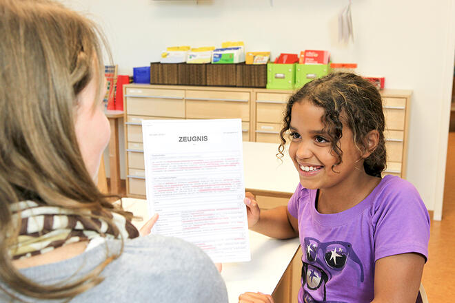 Lächelndes Mädchen zeigt einer Erwachsenen ihr Zeugnis.