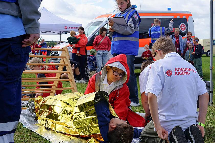 Zwei Kinder der Johanniter versorgen ein verletztes Kind, das auf dem Boden unter eine Rettungsdecke liegt.