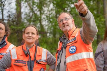 Zwei Johanniter in orangefarbener Einsatzkleidung stehen im Grünen, ein Mann zeigt in die Ferne.