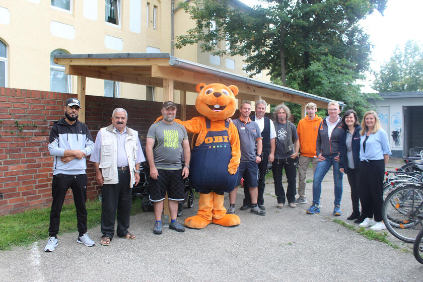 Menschen und ein Obi-Maskottchen vor einem Kinderwagenunterstand