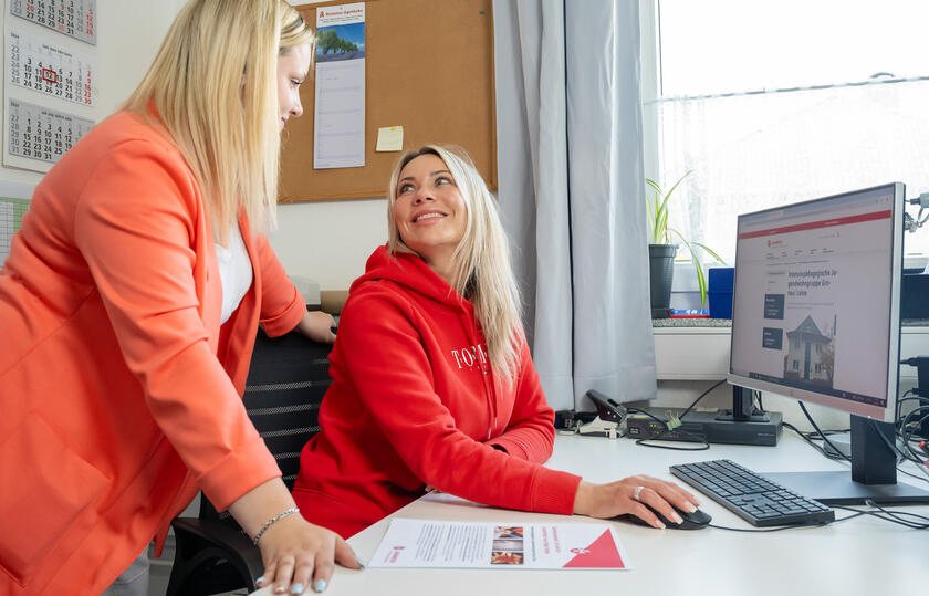 Eine frau sitzt an einem Schreibtisch am Computer und schaut eine andere Frau an, die neben ihr steht. 