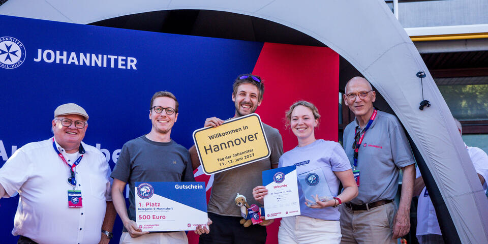 Junge Gewinner*innen präsentieren Urkunde, Gutschein und Scheck vor einem Johanniter‑Messestand.