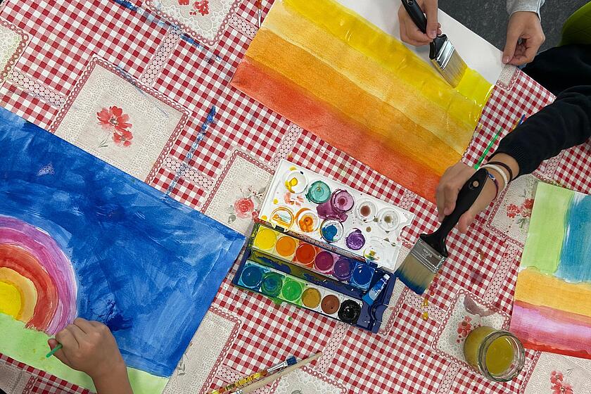 Auf dem Foto sind viele verschiedene Bilder zu erkennen. Die Kinder malen mit dicken Pinseln und Wasserfarbe auf weißte Papierblätter.