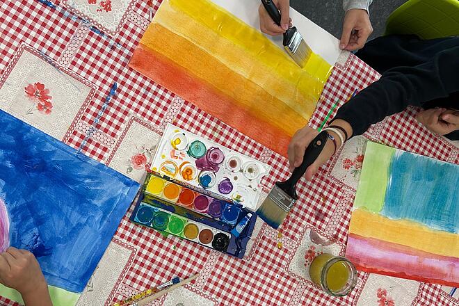 Auf dem Foto sind viele verschiedene Bilder zu erkennen. Die Kinder malen mit dicken Pinseln und Wasserfarbe auf weißte Papierblätter.