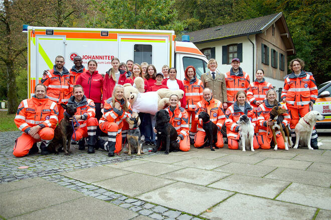Mehrere Johanniter stehen zusammen mit einigen Hunden vor einem Johanniter Einsatzfahrzeug und halten einen großen Teddy in der Hand.
