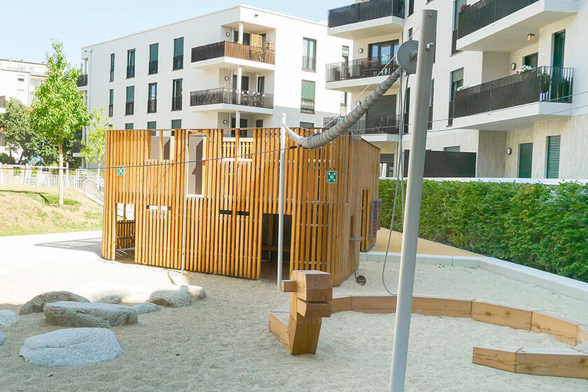 Ein moderner Spielplatz in einer Wohnanlage mit hellen Sandflächen, Holzspielgeräten und einer kleinen Holzhütte, umgeben von mehrstöckigen Wohngebäuden mit Balkonen.