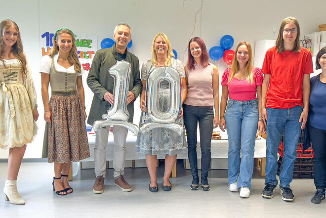 Mehrere Leute stehen vor einem kleinen Buffet. In Hintergrund ist bunte Party Dekoration und zwei Leute halten die Nummer 10 aus Luftballons in der Hand.