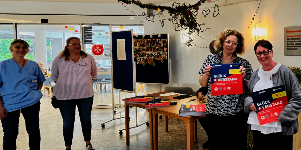vier Personen stehen in einer Halle, zwei davon halten rot-blaue, quadratische Kalnder vor sich