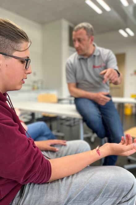 Eine Lehrsituation im Rahmen des Schulsanitätsdienstes: Im Vordergrund sitzt ein Schüler mit einer realistisch geschminkten Wunde am Unterarm. Im Hintergrund erklärt ein Erste-Hilfe-Trainer, was bei einer solchen Verletzung zu tun ist.