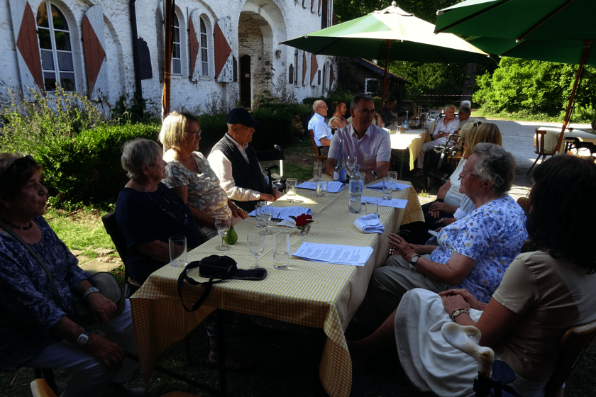 Neun Männer und Frauen in sommerlicher Kleidung sitzen vor einem alte Gutshaus unter einem grünen Sonnenschirm an einem Tisch, auf dem ein gelbweiß kariertes Tischtuch liegt.