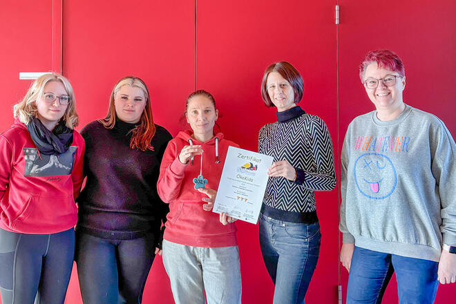 5 Frauen stehen vor einem roten Hintergrund und halten ein Öko-Kids Zertifikat in der Hand.