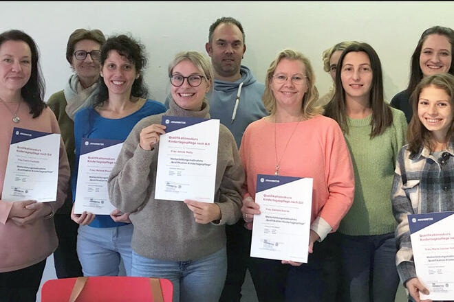 Mehrere Leute lächeln in die Kamera und halten Urkunden für einen Qualifikationskurs Kindertagespflege in der Hand.