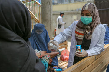 Eine Frau erhält von einer zweiten Frau Desinfektionsmittel, beide tragen einen Mund-Nasen Bedeckung