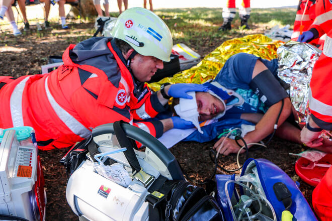 Rettungshelfer*innen stabilisieren im Freien einen verletzten Mann, medizinische Geräte und Notfallausrüstung liegen im Vordergrund.