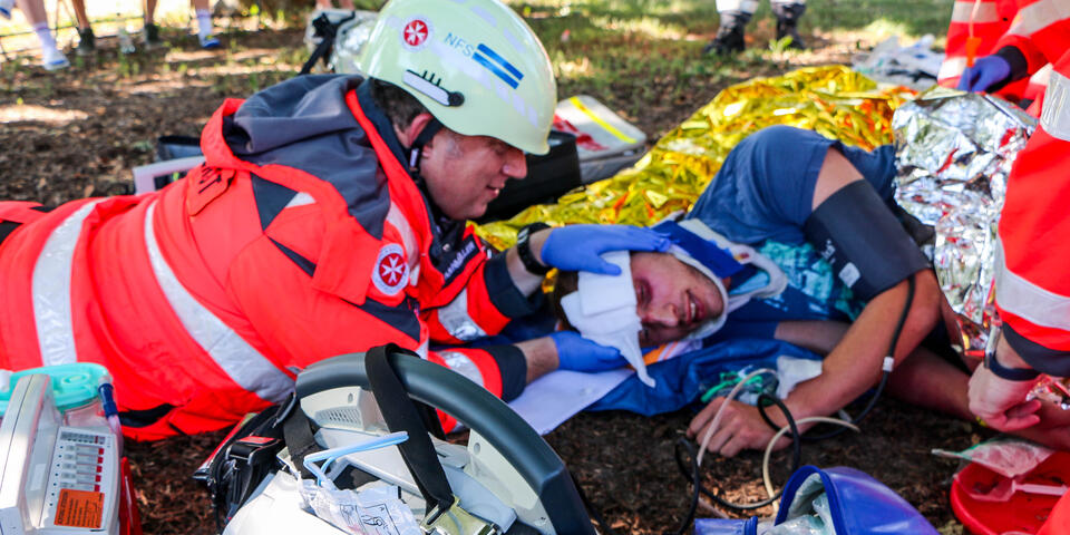 Rettungshelfer*innen stabilisieren im Freien einen verletzten Mann, medizinische Geräte und Notfallausrüstung liegen im Vordergrund.