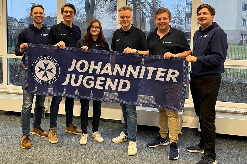 Sechs junge Erwachsene stehen nebeneinander im Johanniter-Jugend-Pullover in einem Raum vor einer Fensterfront. Vor Ihnen ein Banner mit "Johanniter-Jugend".