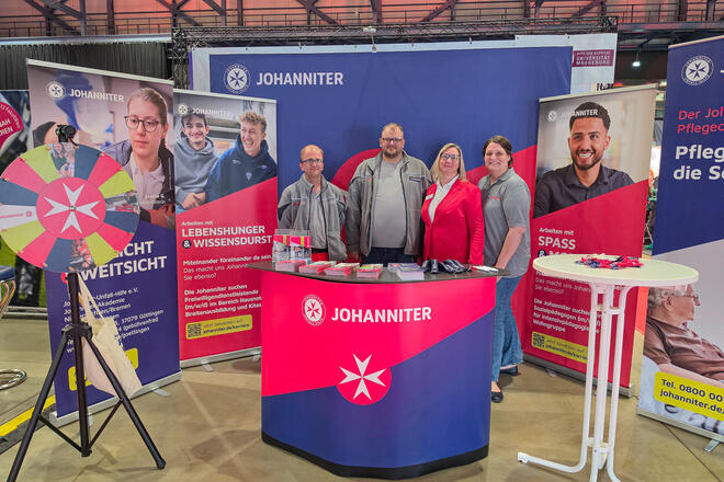 Zwei Frauen und zwei Männer in Johanniter-Kleidung an einem Messestand. Links ist ein Glücksrad zu sehen.