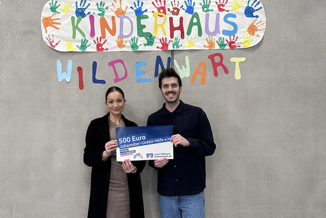 Charlotte Pieslinger De Bernardin übergibt einen Spendenscheck an Einrichtungsleiter Johannes kern vor einer Grauen Wand mit einem Schild der Einrichtung umrahmt von bunten Handabdrücken.