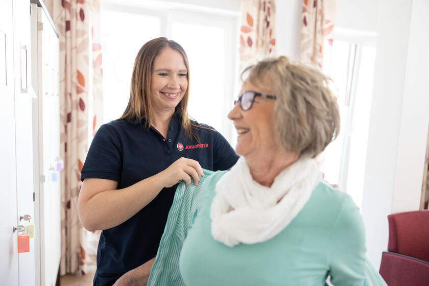 Johanniter-Mitarbeiterin hilft Seniorin beim Anziehen, beide lächeln
