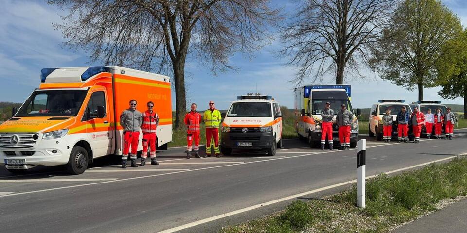 Zwei Rettungswagen, ein Notarzteinsatzfahrzeug und zwei Mannschaftstransportwagen stehen aufgereiht auf einer Landstraße Menschen in Einsatzkleidung stehen vor den Fahrzeugen.