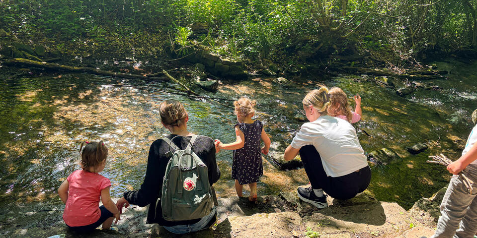 Erwachsene und Kleinkinder knieen an einem Bach in einem Wald.