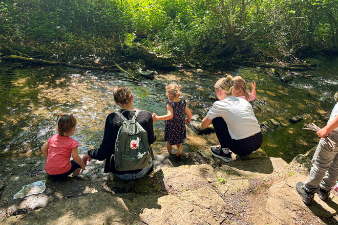 Erwachsene und Kleinkinder knieen an einem Bach in einem Wald.