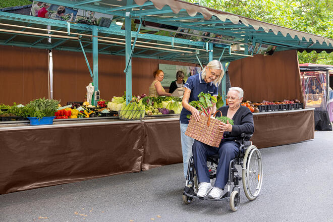 Eine Frau überreicht einer älteren Frau, die in einem Rollstuhl sitzt, einen Korb mit Gemüse. Sie befinden sich auf einem Wochenmarkt.