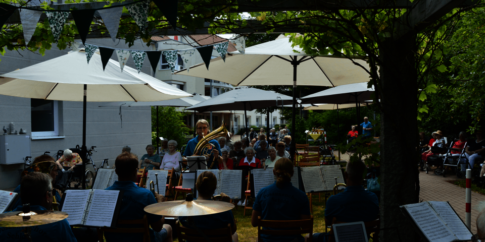 Unter weißen Sonnenschirmen sitzen Zuhörende in mehreren Reihen. Im Vordergrund ist der in türkisfarbene Poloshirts gekleidete Blaskapelle zu sehen.