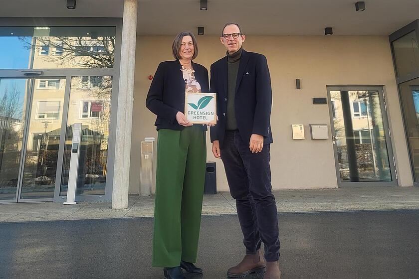 Hotelleitung Helga Butendeich und Auditor Roberto Pareras halten GreenSign Zertifikat vor dem INCLUDiO Hotel