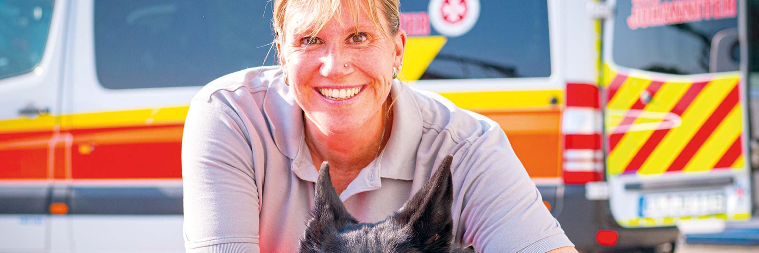 Einsatzkraft der Johanniter sitzt lachend auf dem Boden und hält einen schwarzen Hund, der fröhlich die Zunge herausstreckt. Im Hintergrund steht ein Einsatzfahrzeug