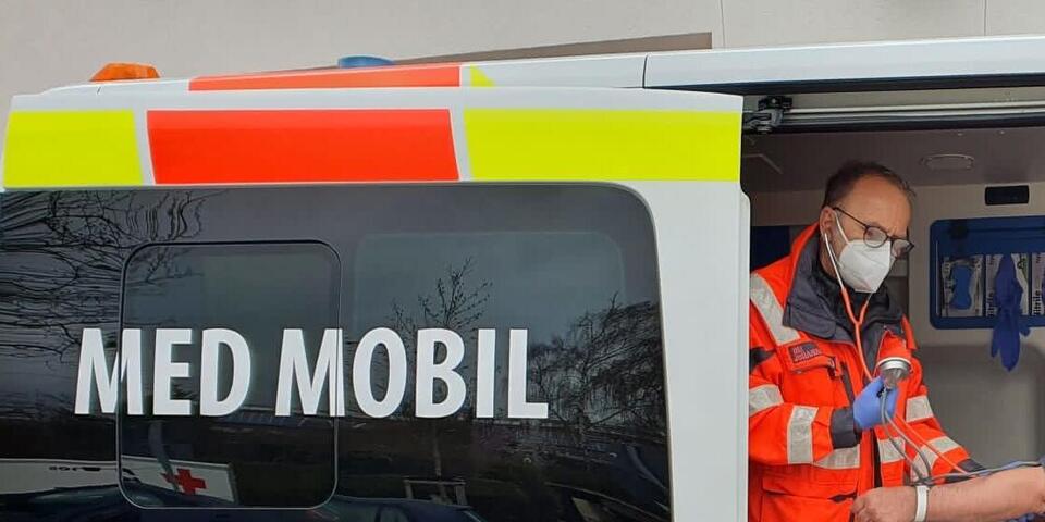 Einsatzkraft im „MED MOBIL“ untersucht eine Person, davor steht jemand in lilafarbener Johanniter-Jacke.