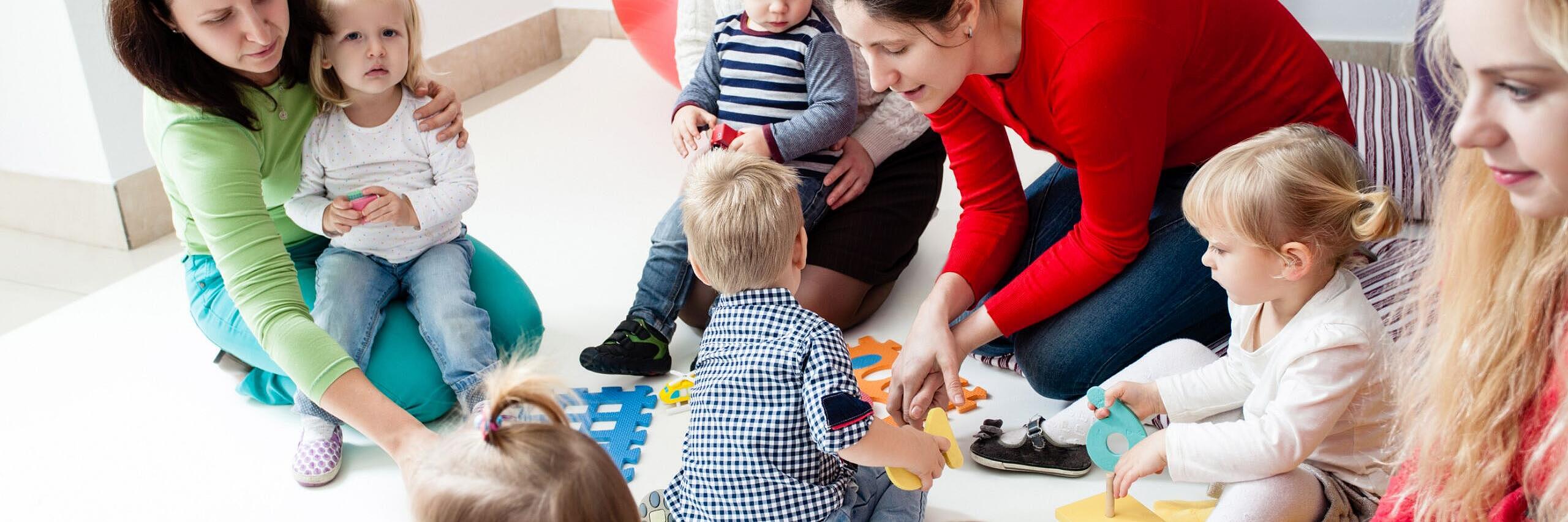 Vier Erzieherinnen und 6 kleine Kinder sitzen zusammen am Boden. Um sie herum liegt Spielzeug, mit welchem sie gerade spielen und zwei der Kinder sitzt auf dem Schoß zwei Erzieherinnen.