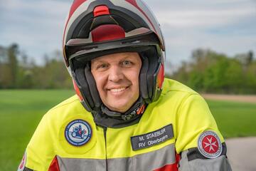 Johanniter im gelben Motorrad-Einsatzanzug mit Helm lächelt in die Kamera, im Hintergrund Wiese.